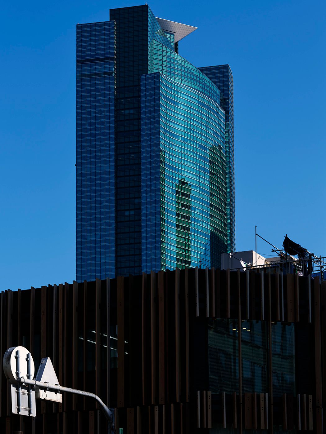 Modern glass skyscraper with teal reflective facade against clear blue sky, with dark slatted building in foreground.