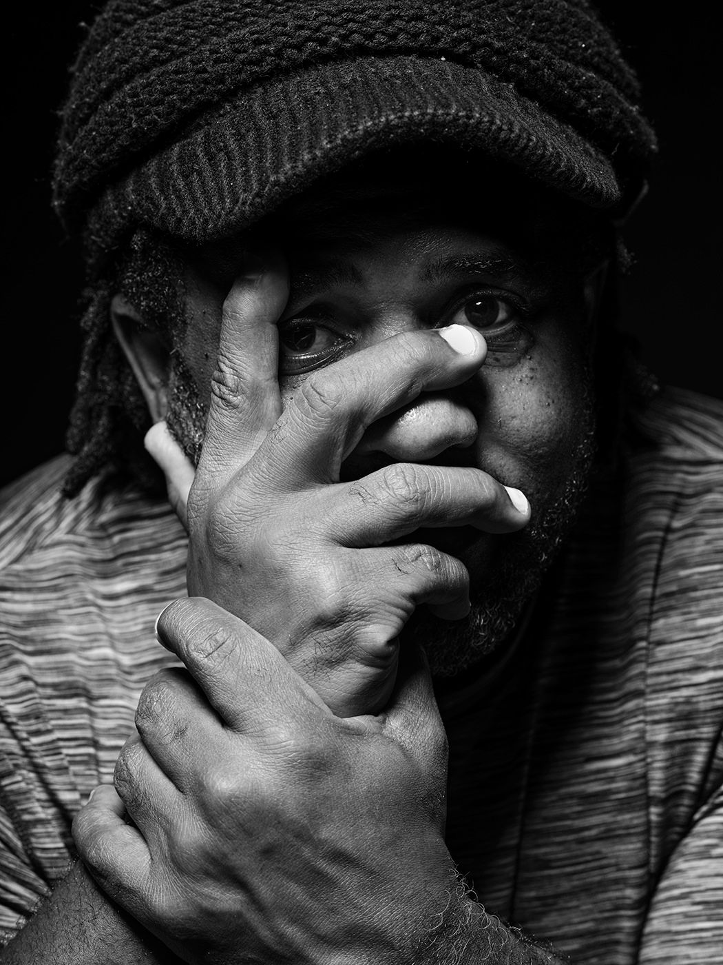 Black and white portrait of a man in a flat cap covering his face with his hand, dramatic studio lighting. Victor Wooten, musician, bass.