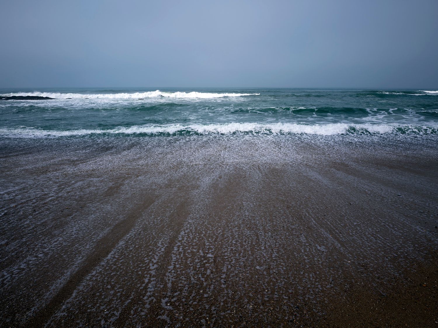 Stormy ocean waves crashing on a sandy beach under overcast skies with turquoise water and seafoam.
