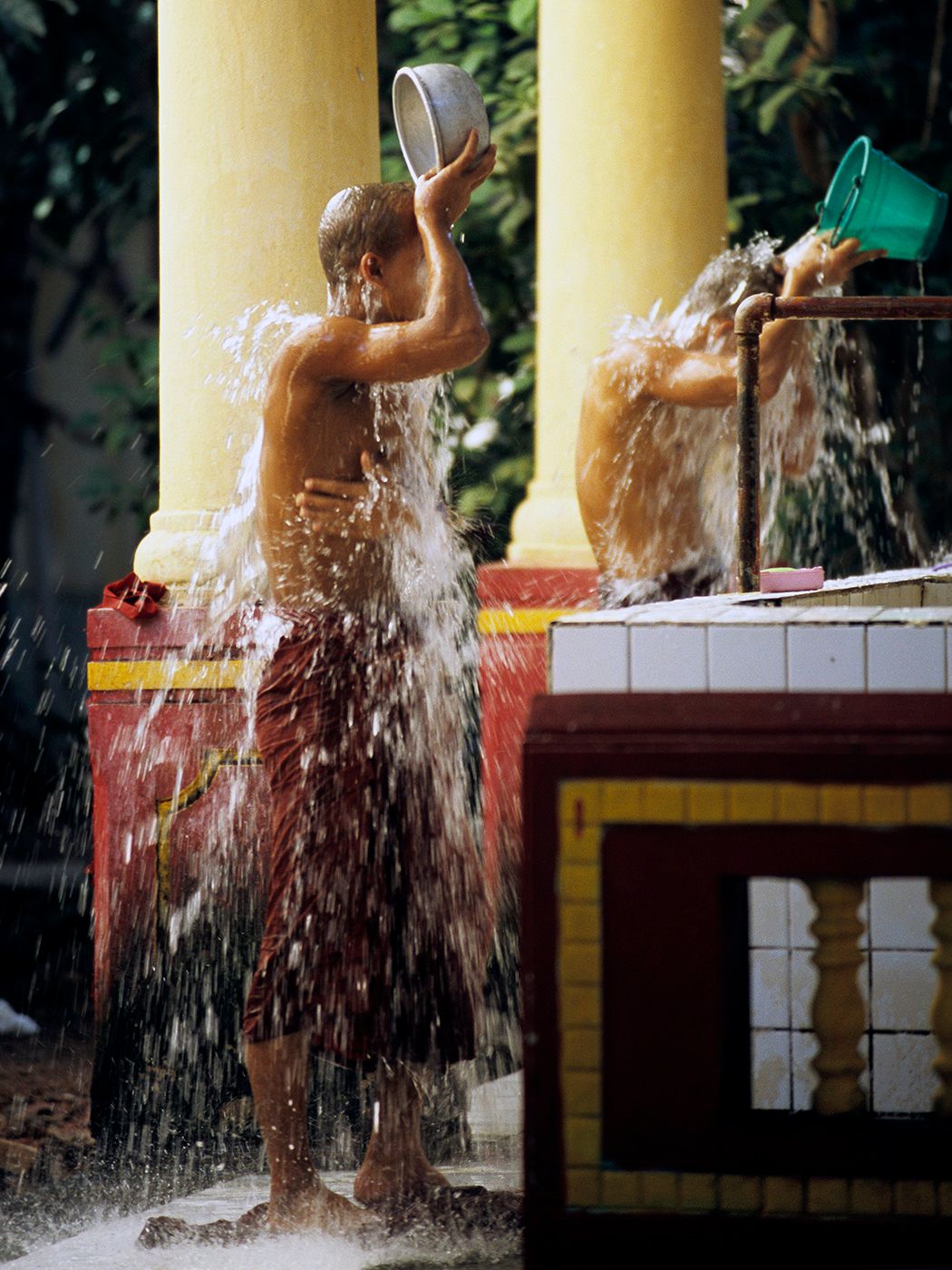 Two monks bathing outdoors by pouring buckets of water over themselves near colorful temple columns.