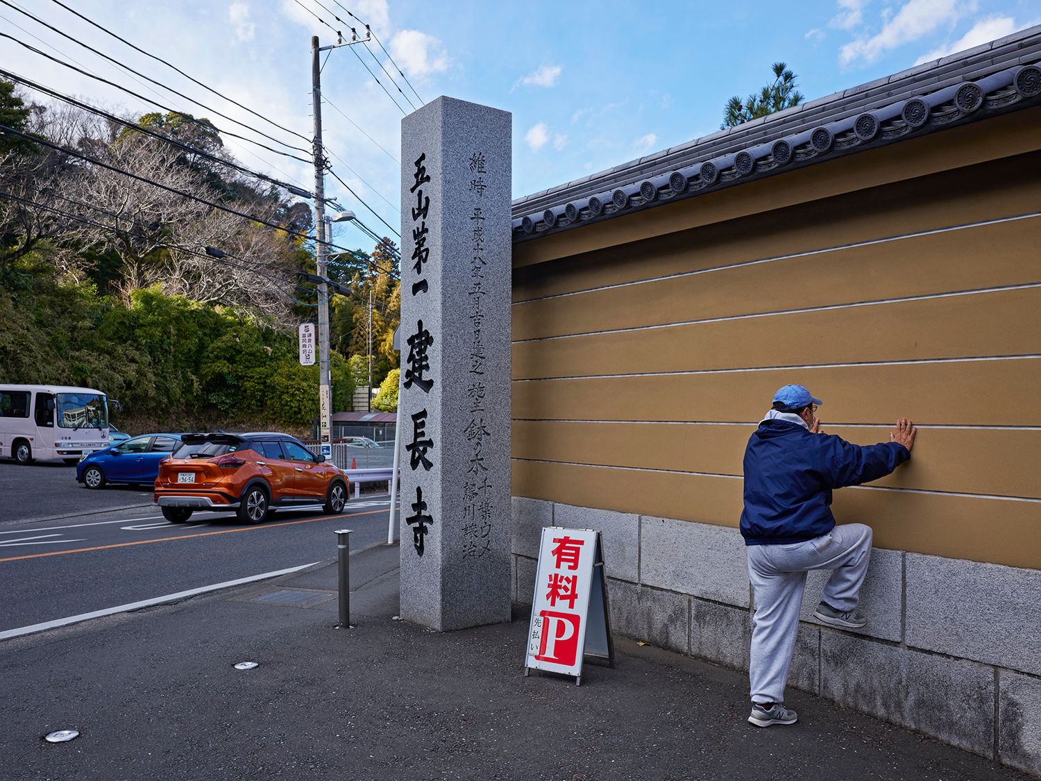 A person examines a Japanese stone monument near a temple wall with a paid parking sign in Kyoto, Japan.