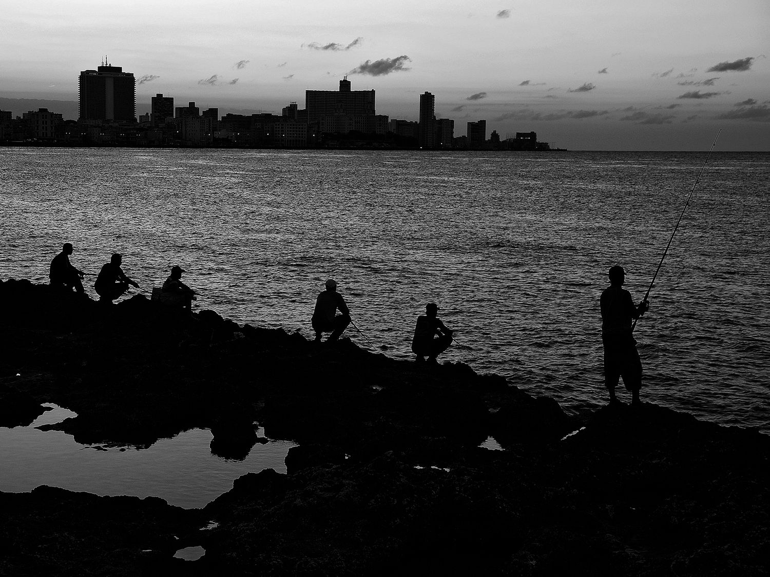 Silhouettes of fishermen on rocky shoreline at dusk with city skyline reflected on calm water in black and white.