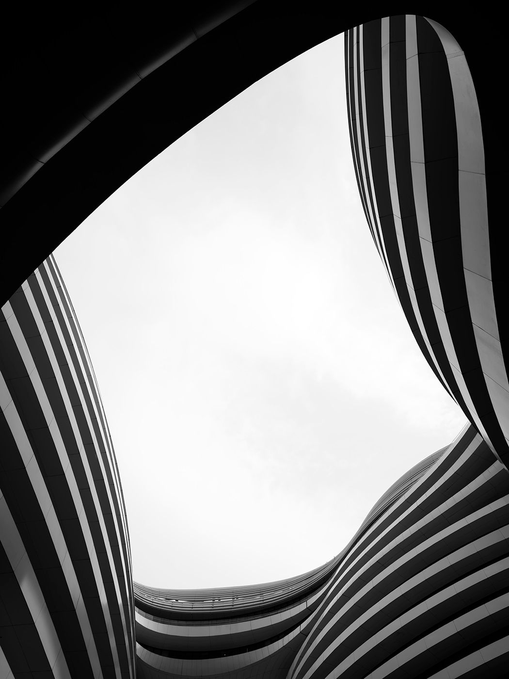 Black and white architectural photo looking up through curved striped building openings toward a bright sky.