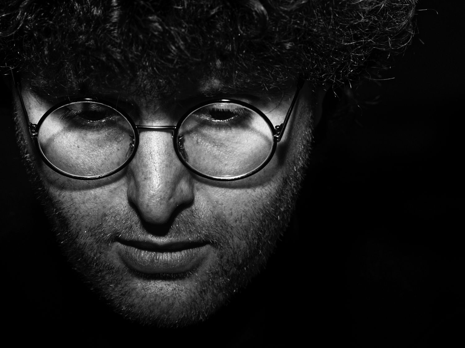 Black and white close-up portrait of a man with curly hair and round glasses in dramatic low-key lighting. Bornai Tibor