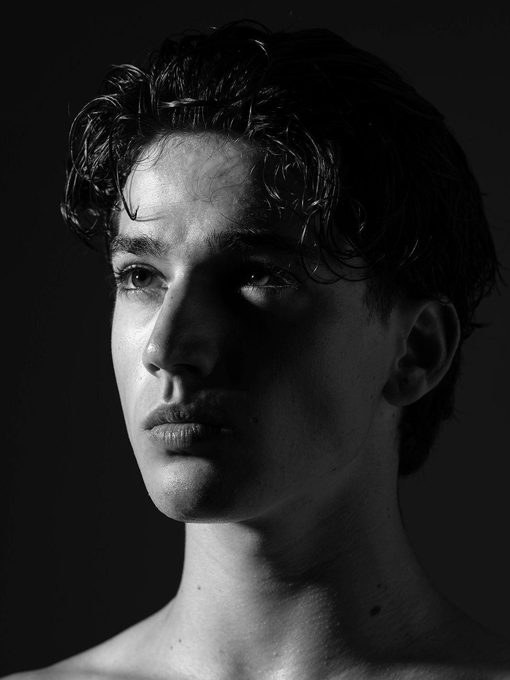 Black and white portrait of a young man with curly dark hair, shirtless, against a dark background.