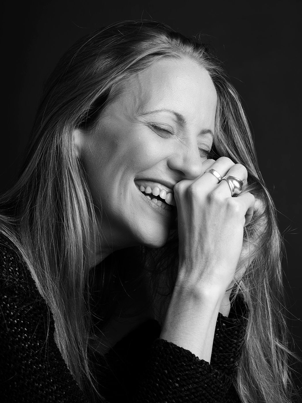 A laughing blonde woman covers her mouth with ringed hands in a black and white portrait.