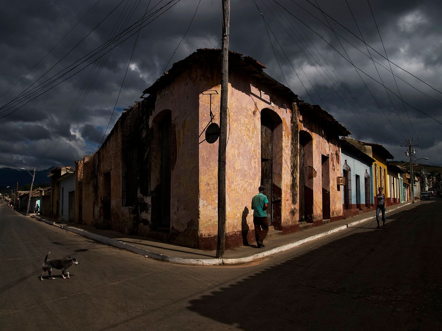 A person walks past a weathered corner building under dramatic storm clouds on a quiet Latin American street.