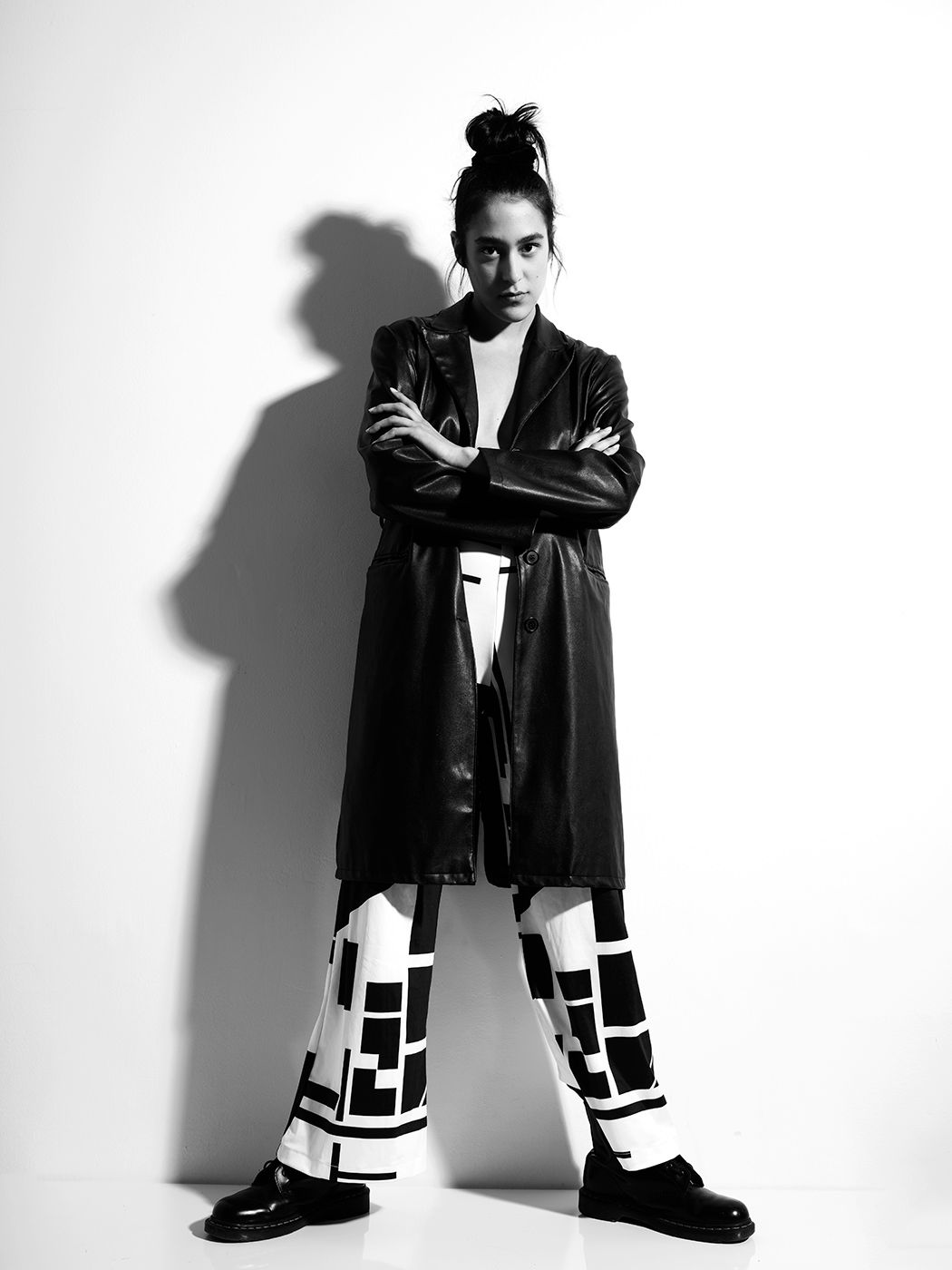 Black and white photo of a model in a leather coat and geometric patterned pants, arms crossed, studio background.