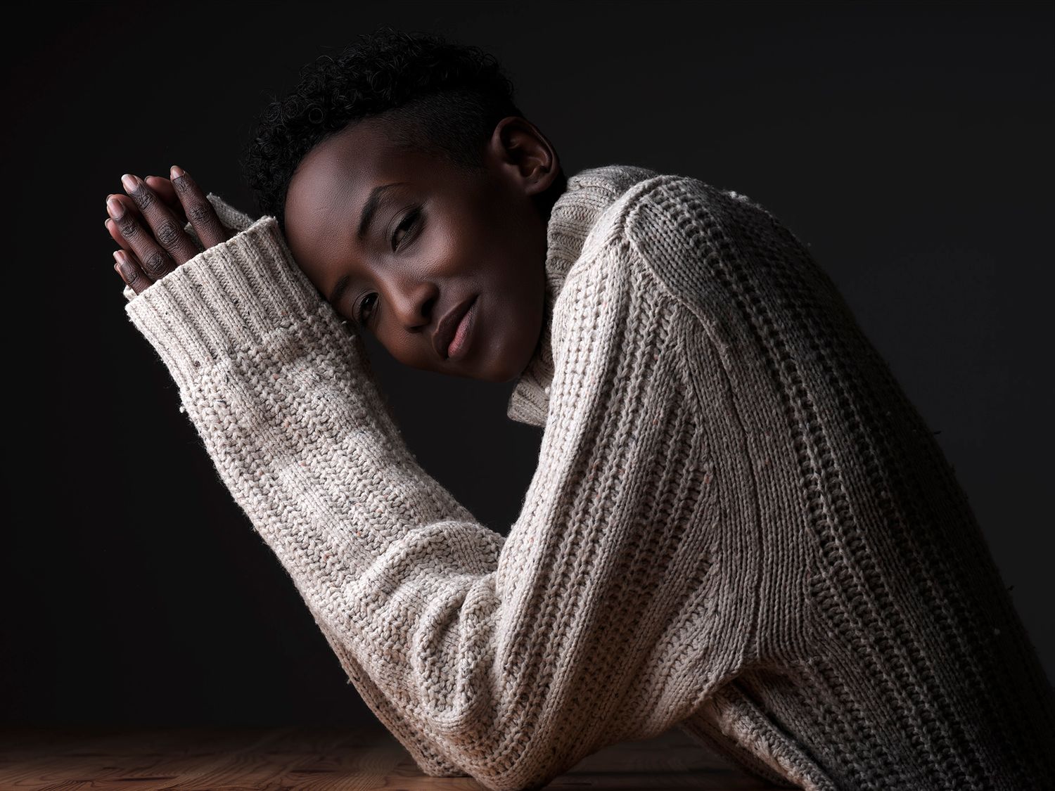 Young Black woman in cream cable-knit sweater rests head on arms, gazing softly in moody studio portrait.