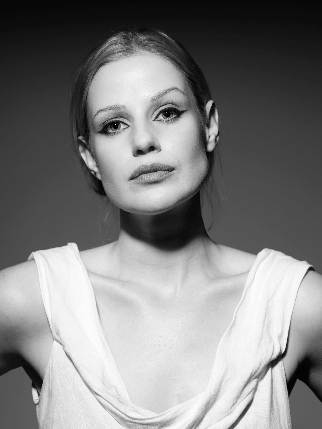 Black and white portrait of a young woman with pulled-back hair, dramatic eye makeup, wearing a white sleeveless top.