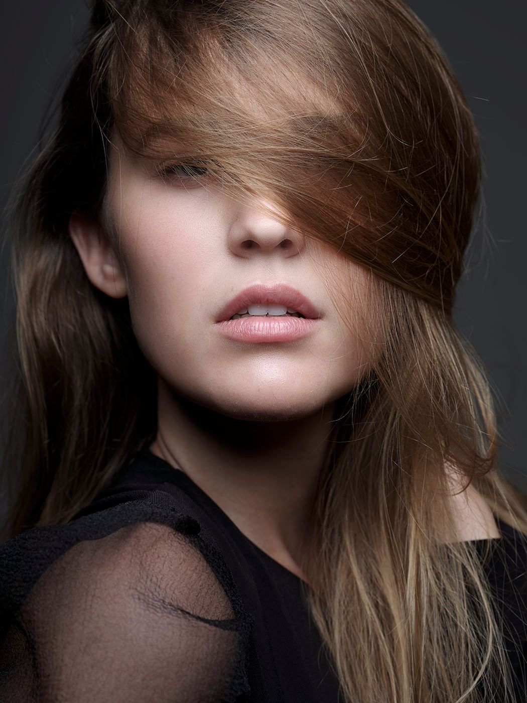 A young woman with long brown hair blowing across her face, wearing a black sheer top against a dark background.