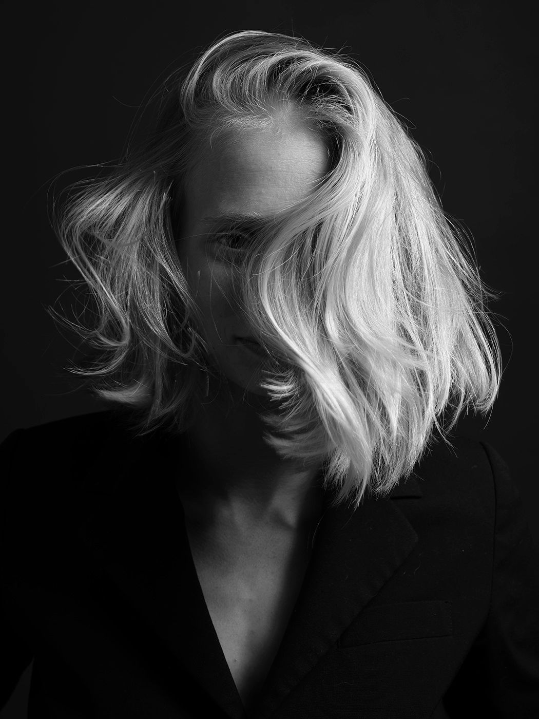 Black and white portrait of a blonde woman with tousled hair wearing a dark blazer against a dark background.