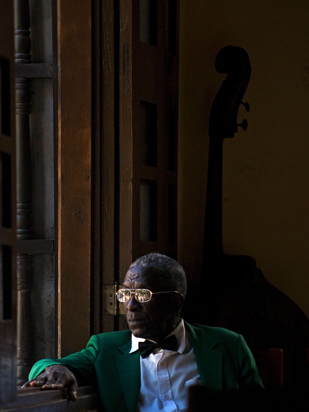 Elderly jazz musician in green blazer and bow tie sits by window with double bass in dim light.