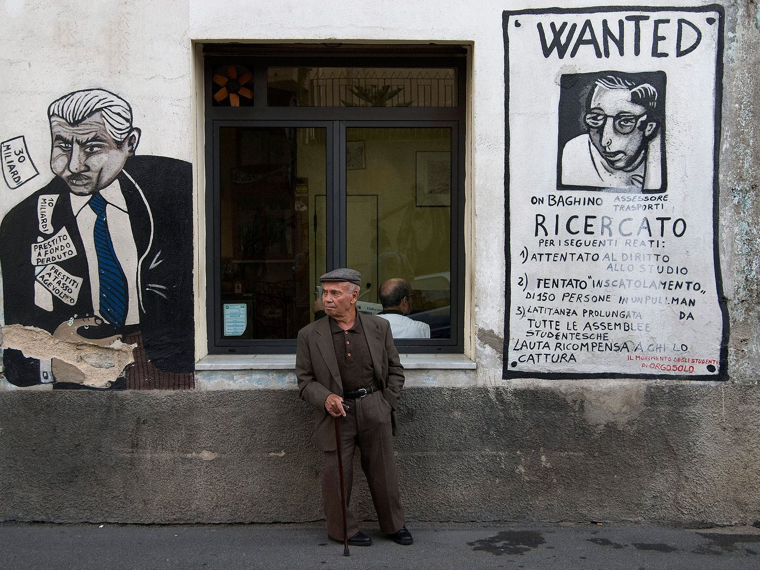 Elderly man with cane stands beside Italian street art mural and a 'Wanted' poster on a weathered building wall.