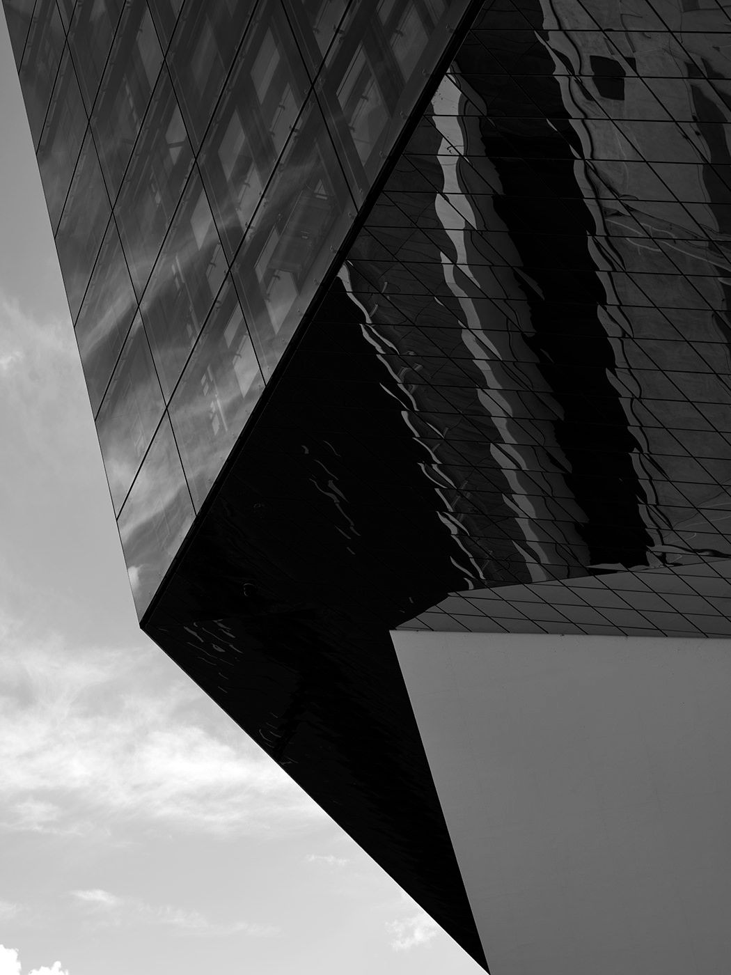 Black and white architectural photo of a modern glass building with dramatic angular geometry and cloud reflections.
