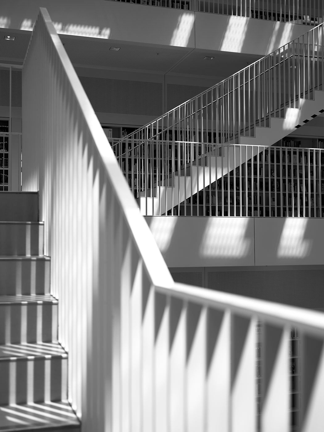 Black and white photo of modern building interior staircase with geometric railings and dramatic light and shadow patterns.
