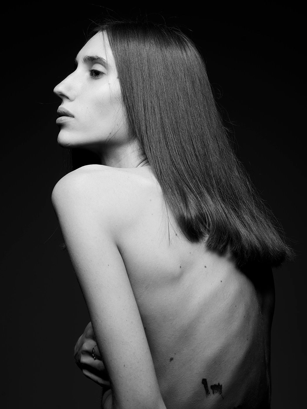 Black and white portrait of a woman with long straight hair, bare back turned to camera, arms crossed.