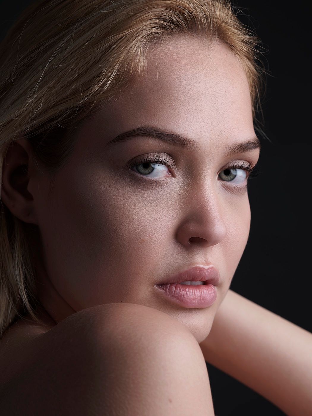 Close-up portrait of a young woman with blonde hair, natural makeup, and glowing skin against a dark background.