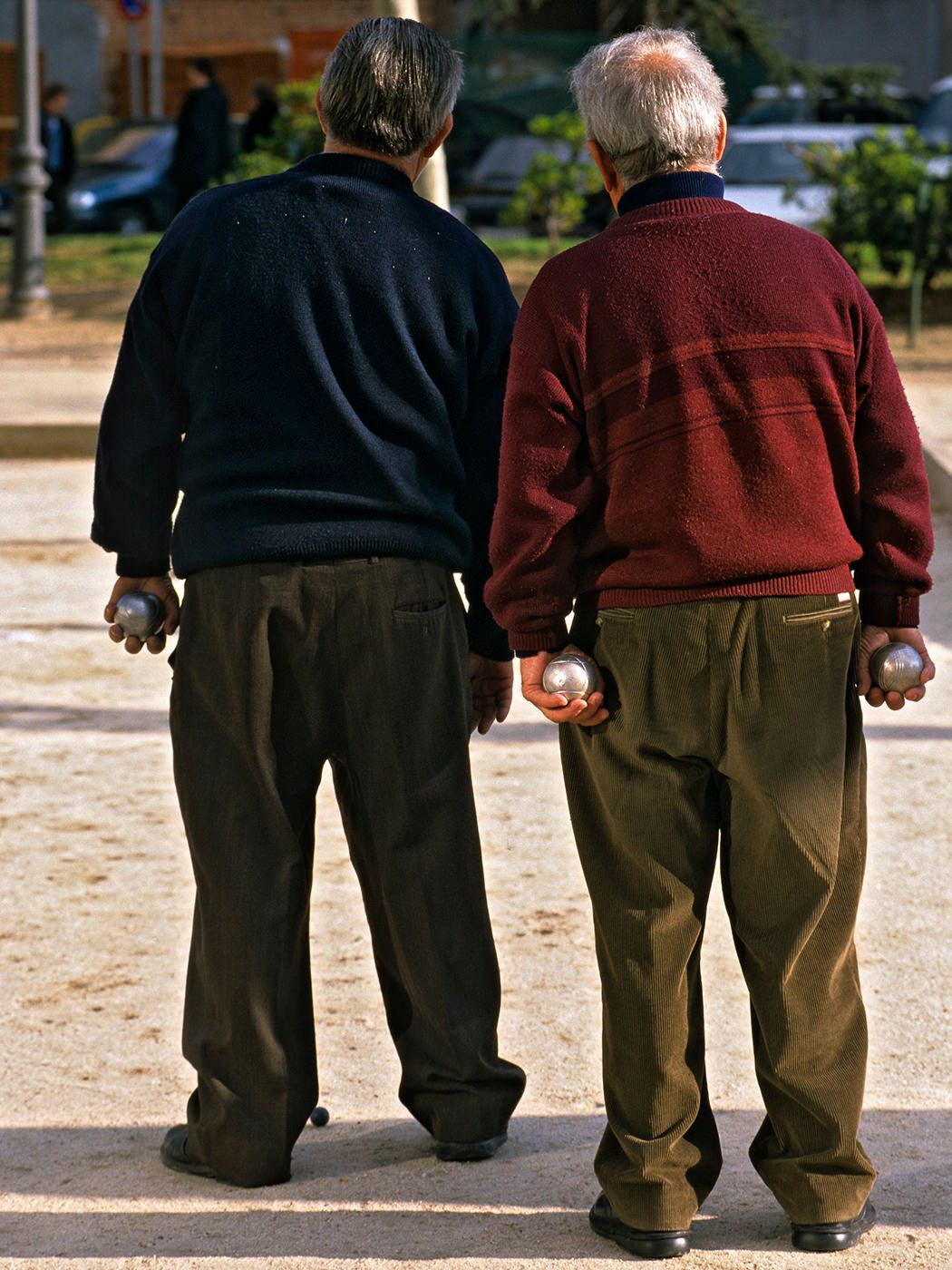 Two elderly men playing petanque, viewed from behind, holding metal boules balls in an outdoor court.