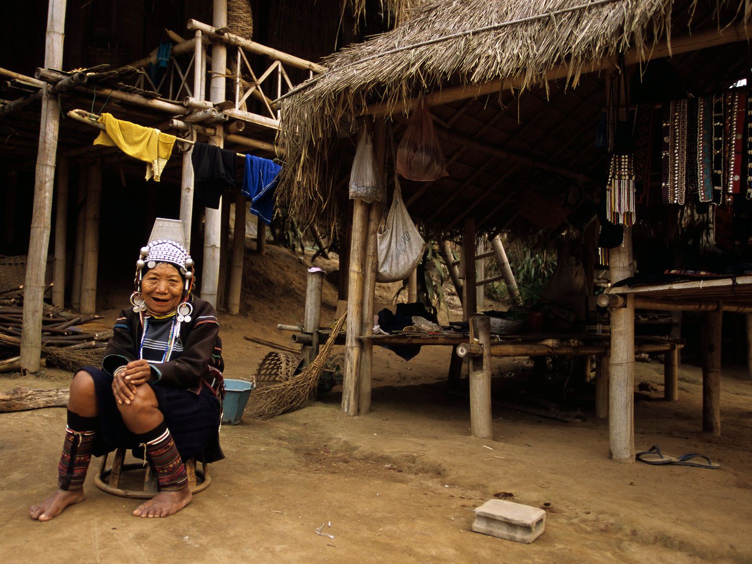 Elderly Kayan tribe woman wearing traditional neck rings sits outside a thatched stilt house in a rural Southeast Asian village.