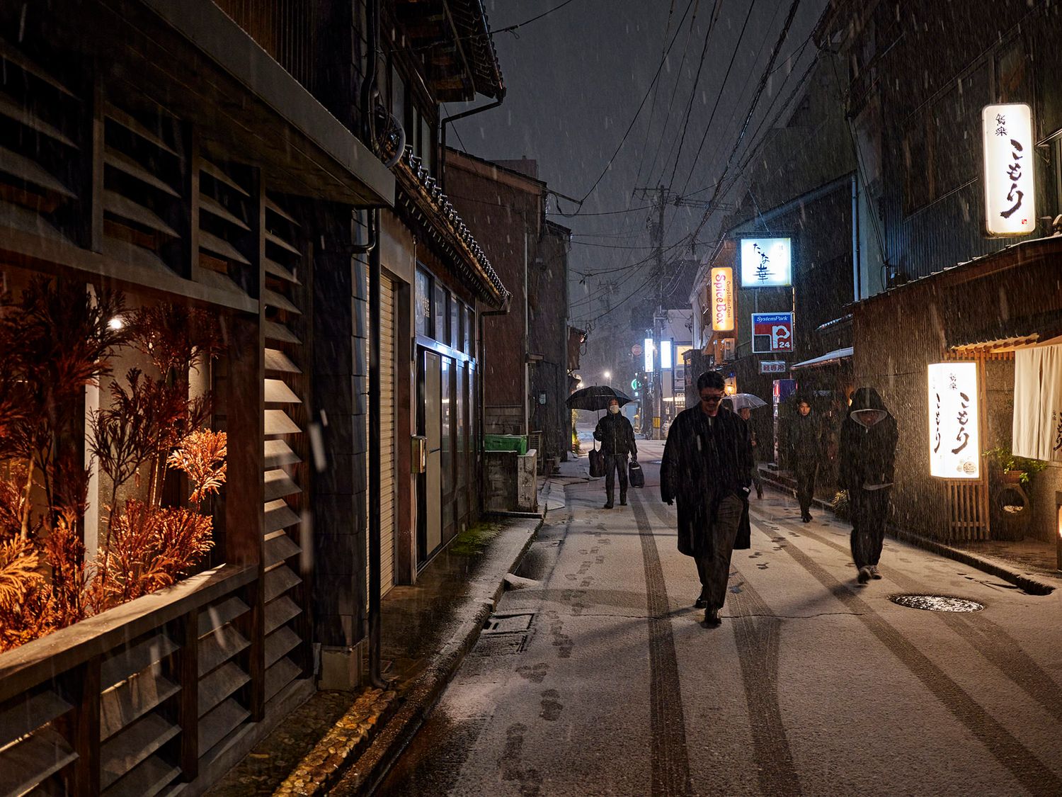 Pedestrians walk along a quiet Japanese alley at night, with warm glowing storefronts and light snow falling.