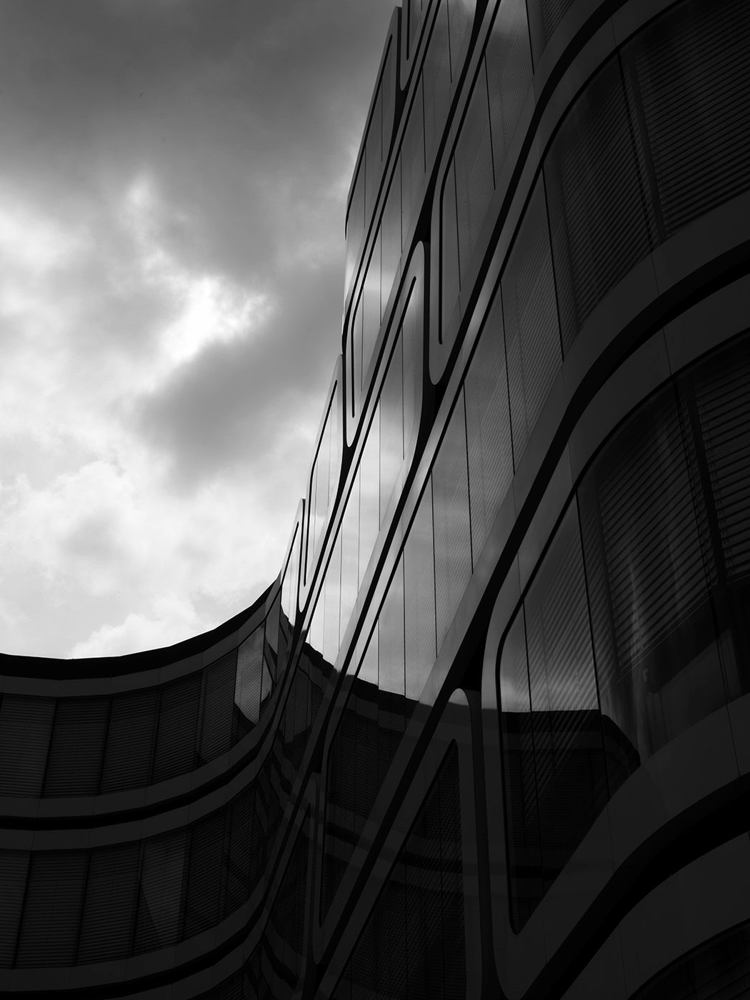 Black and white photo of modern glass building with curved architecture against dramatic cloudy sky.