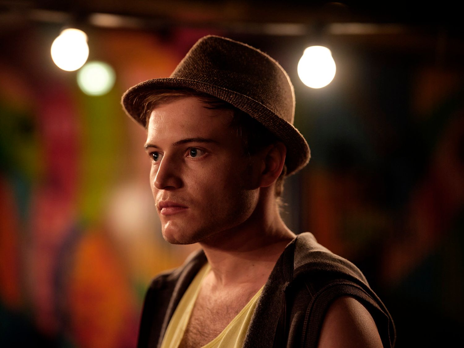 Young man wearing a fedora hat standing under stage lights at a colorful venue.