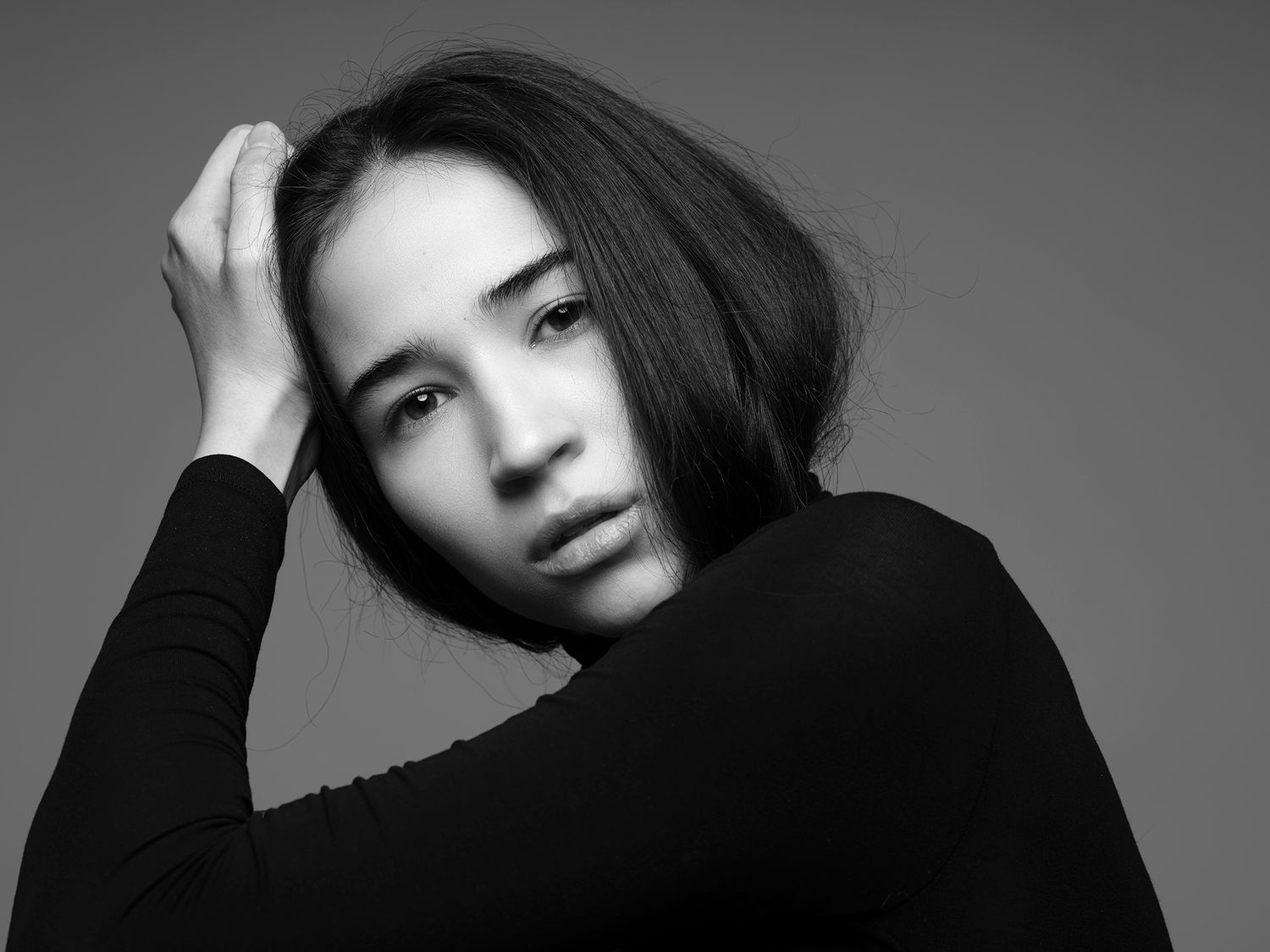Black and white portrait of a young woman in a black turtleneck, hand in hair, against a gray background.