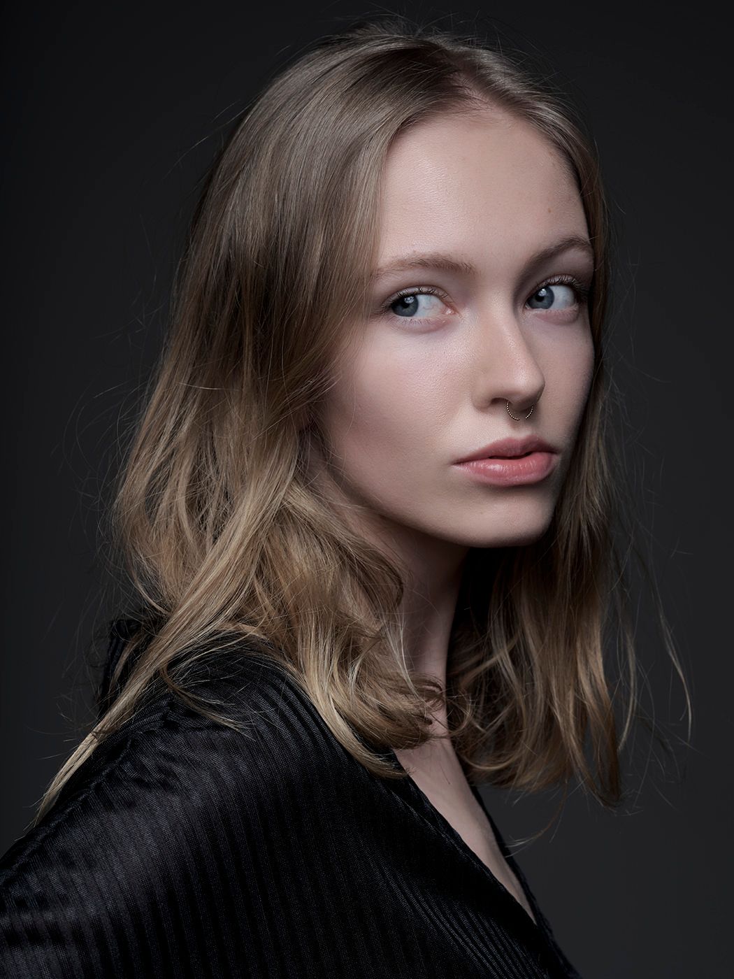 Young woman with blonde hair wearing a black jacket poses against a dark studio background.