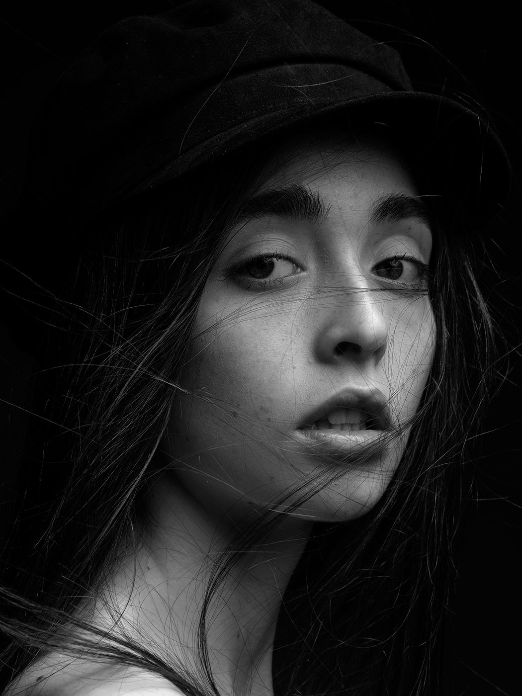 Black and white portrait of a young woman wearing a dark hat with hair blowing across her face against a dark background.