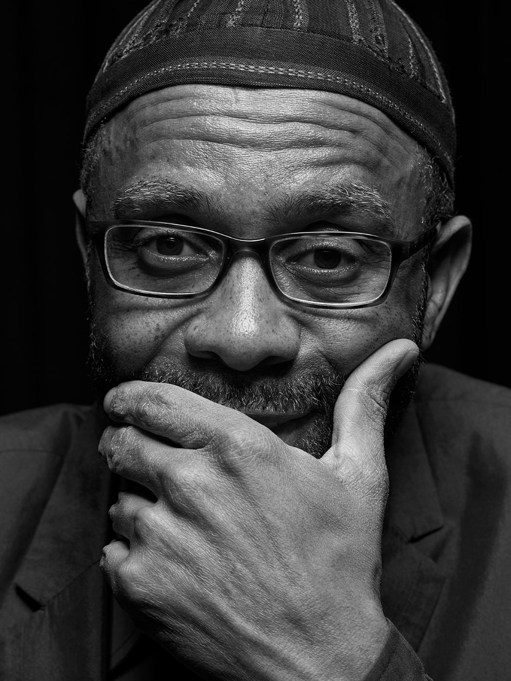 Black and white portrait of a thoughtful man with glasses and kufi cap, hand raised to chin. Kenny Garrett
