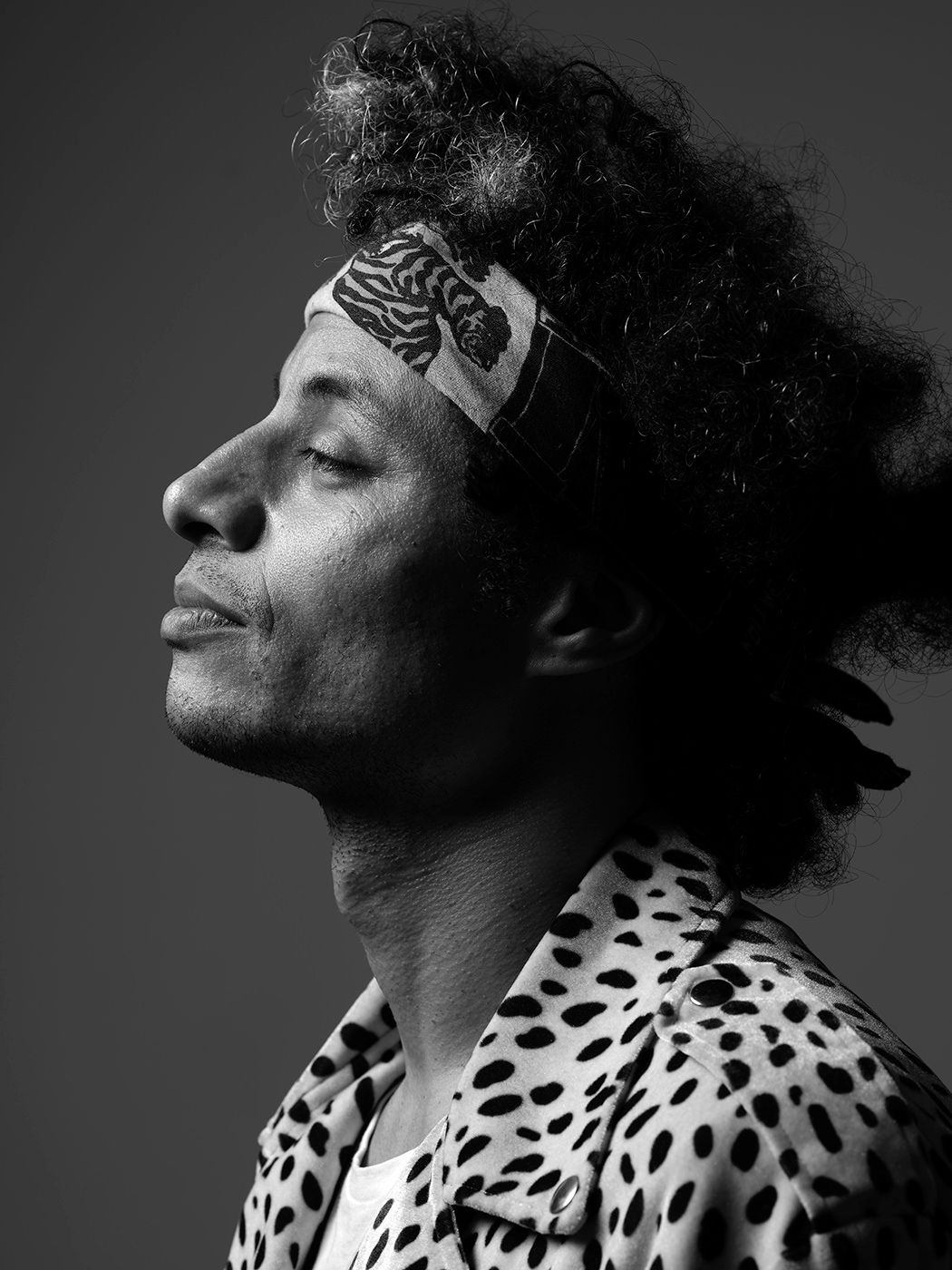 Black and white profile portrait of a man with curly hair wearing a patterned headband and dalmatian print shirt. José James