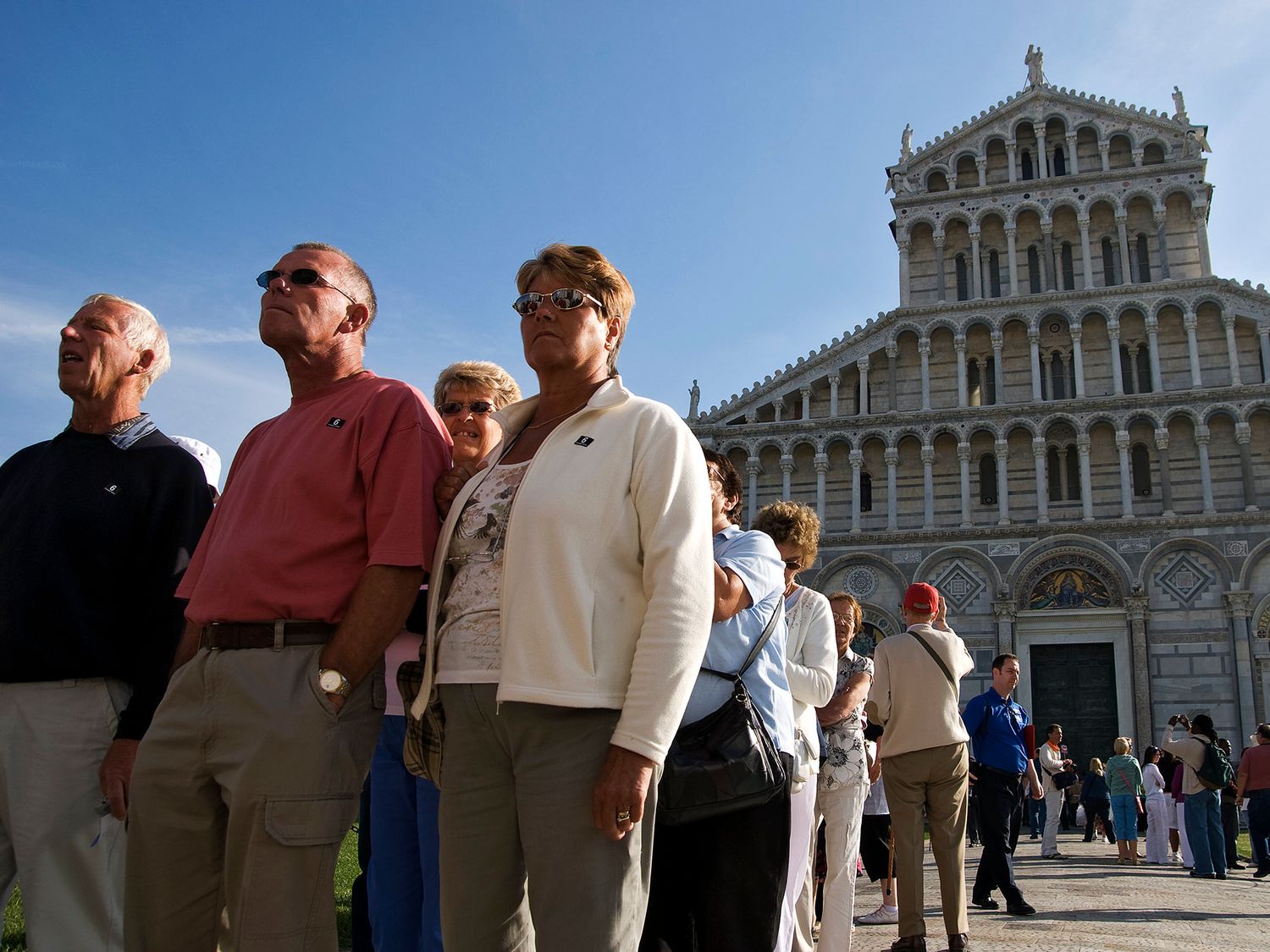 Tourists queue outside the ornate Pisa Cathedral in Italy under a clear blue sky.