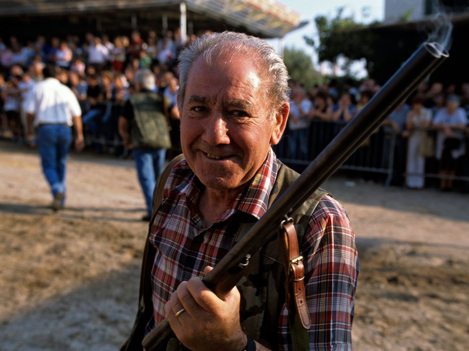 Elderly man in plaid shirt holding a rifle at an outdoor event with crowd in background.