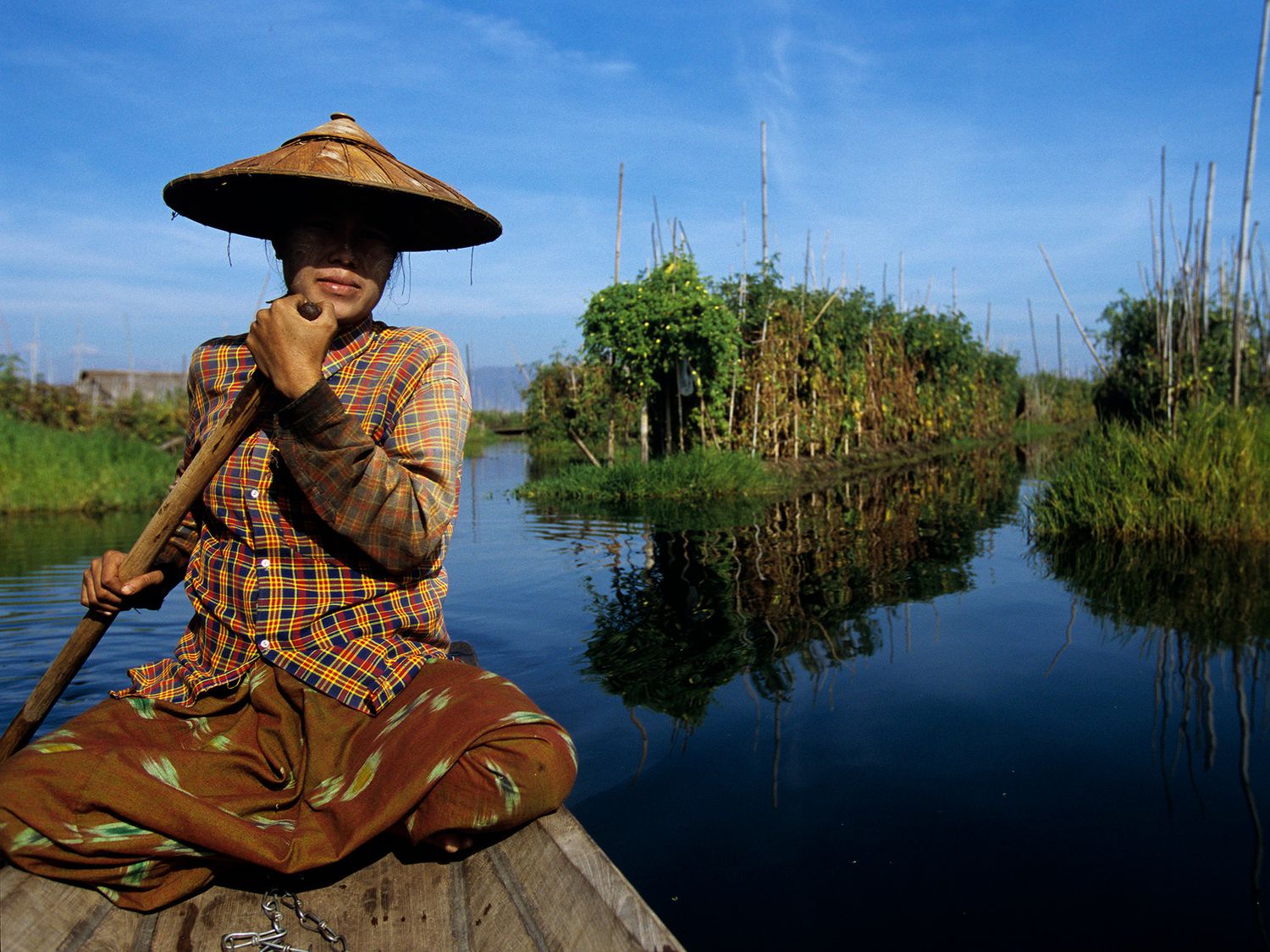A woman in a traditional hat rows a wooden boat through a calm river surrounded by lush greenery in Myanmar.