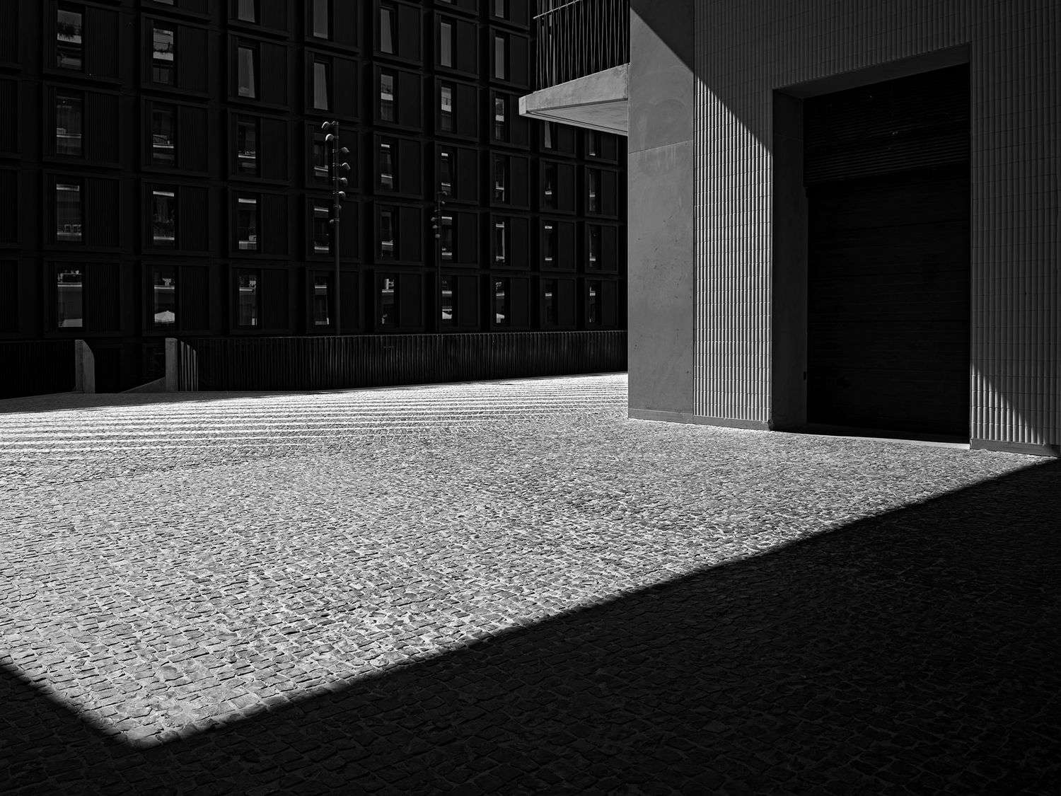 Black and white photo of a sunlit cobblestone plaza beside a modern building with dramatic shadows.
