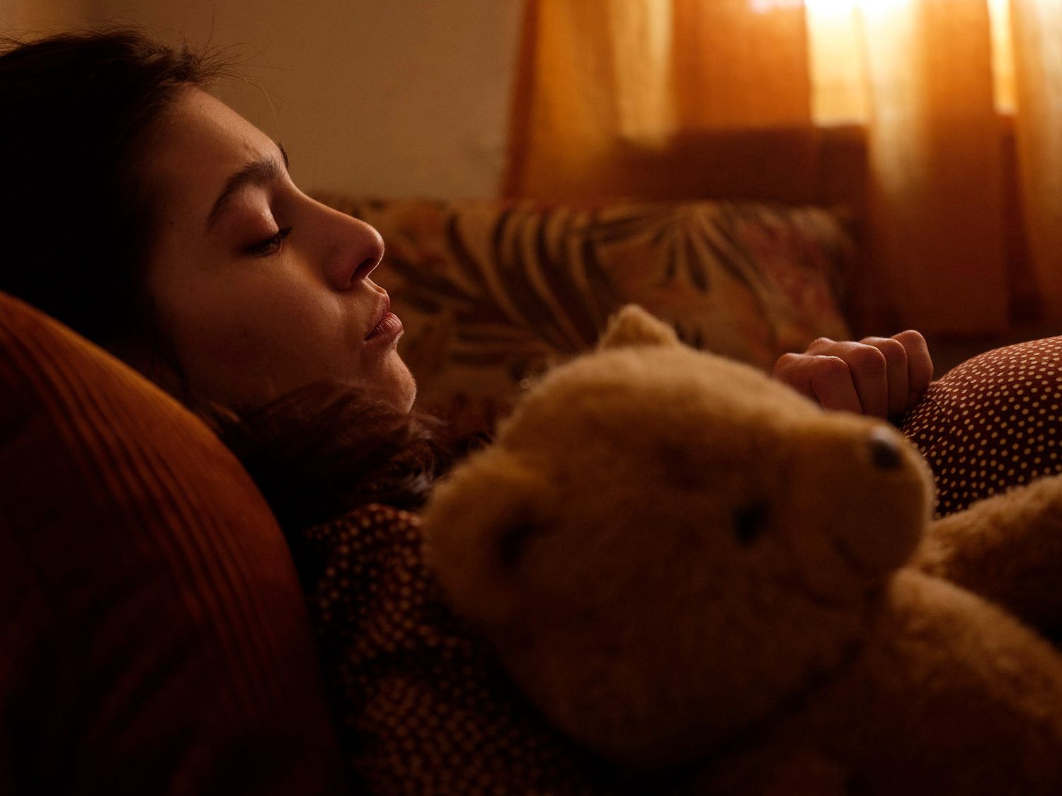 Young woman sleeping peacefully on a couch with a teddy bear in warm golden light.