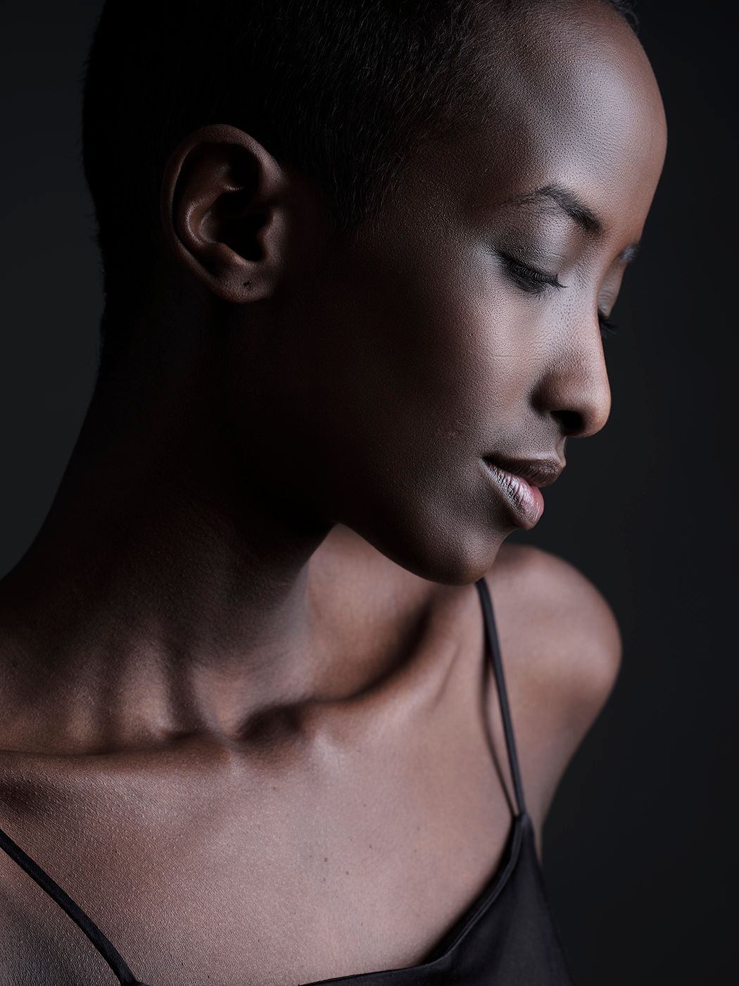 A bald Black woman with closed eyes poses gracefully against a dark background in a sleek spaghetti-strap top.