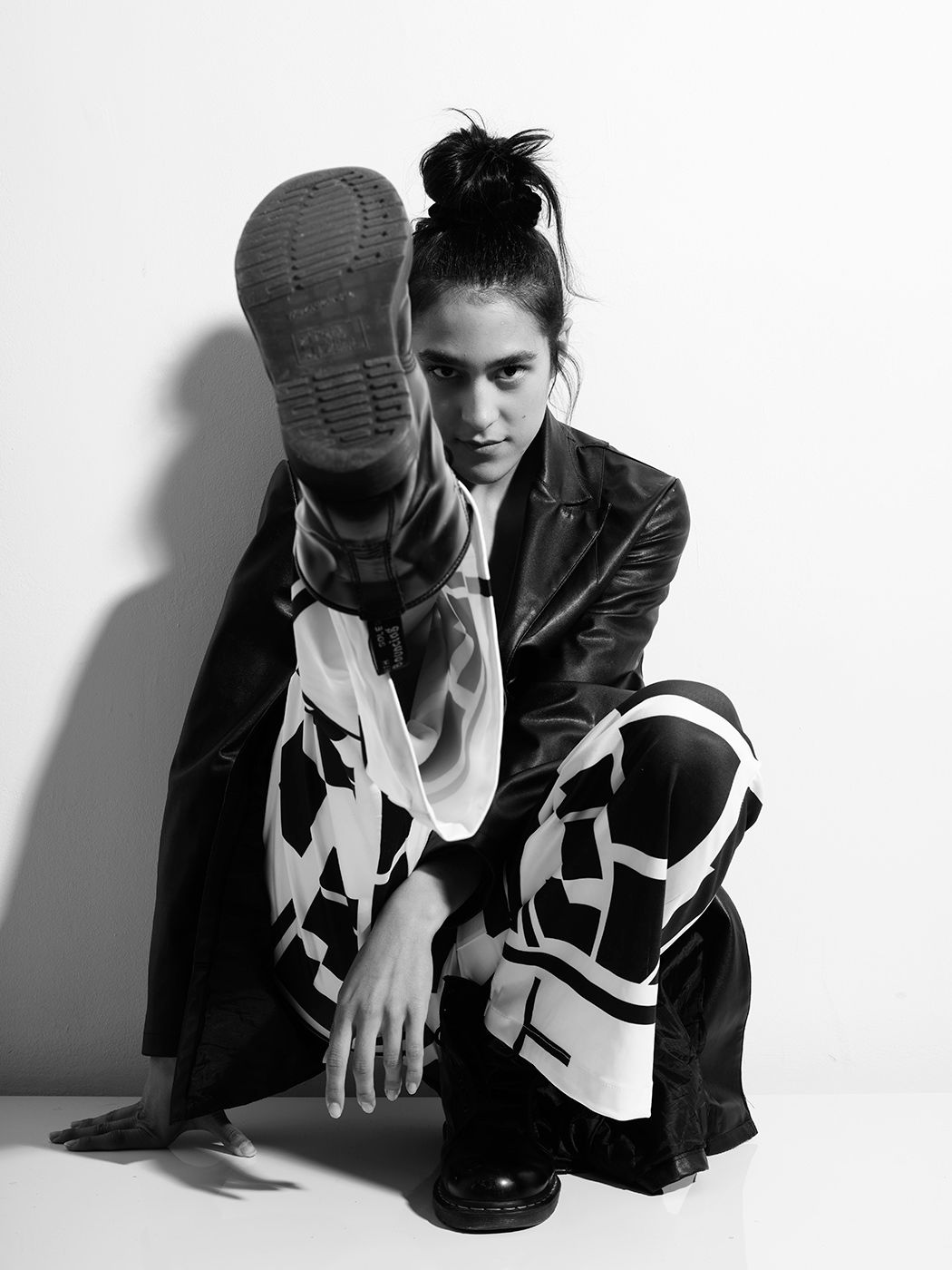 Black and white photo of a model crouching in bold graphic pants, leather jacket, and boots with one leg raised.