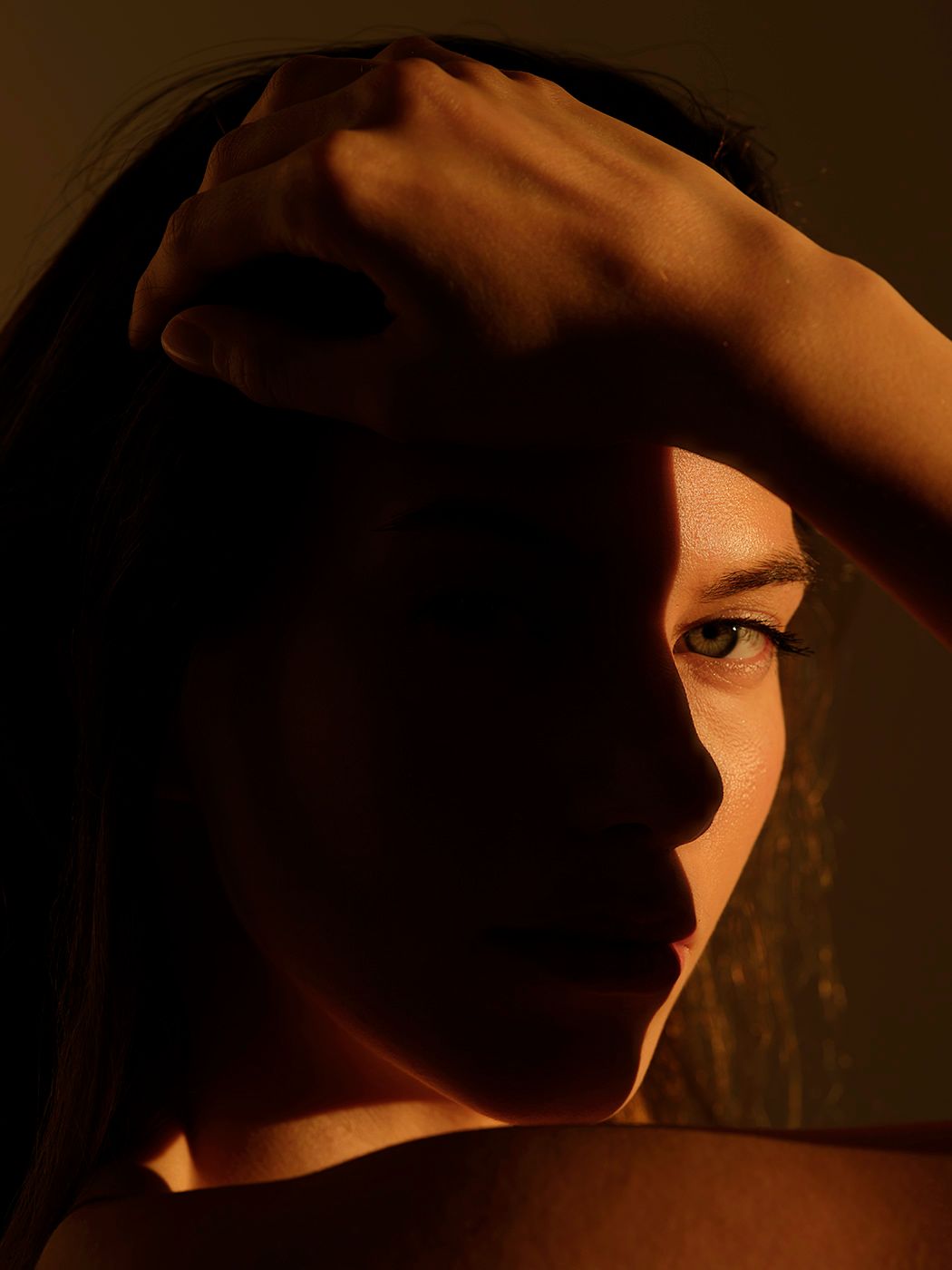 Close-up portrait of a person with dramatic shadow lighting, hand raised near head against dark background.