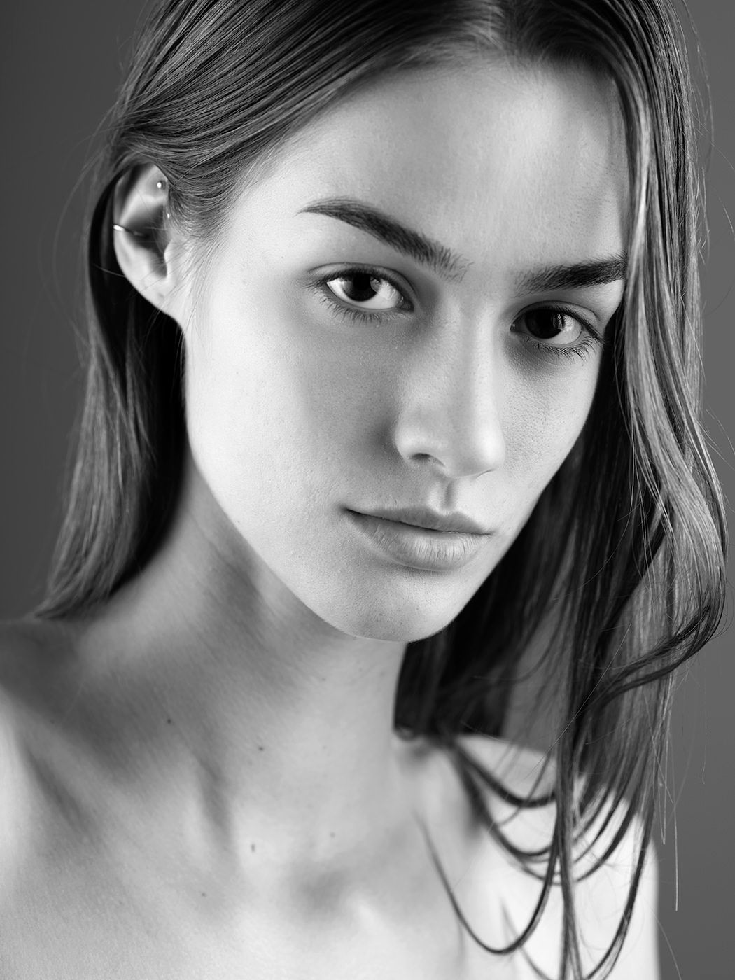 Black and white portrait of a young woman with long hair, natural makeup, and a nose piercing, gazing at the camera.
