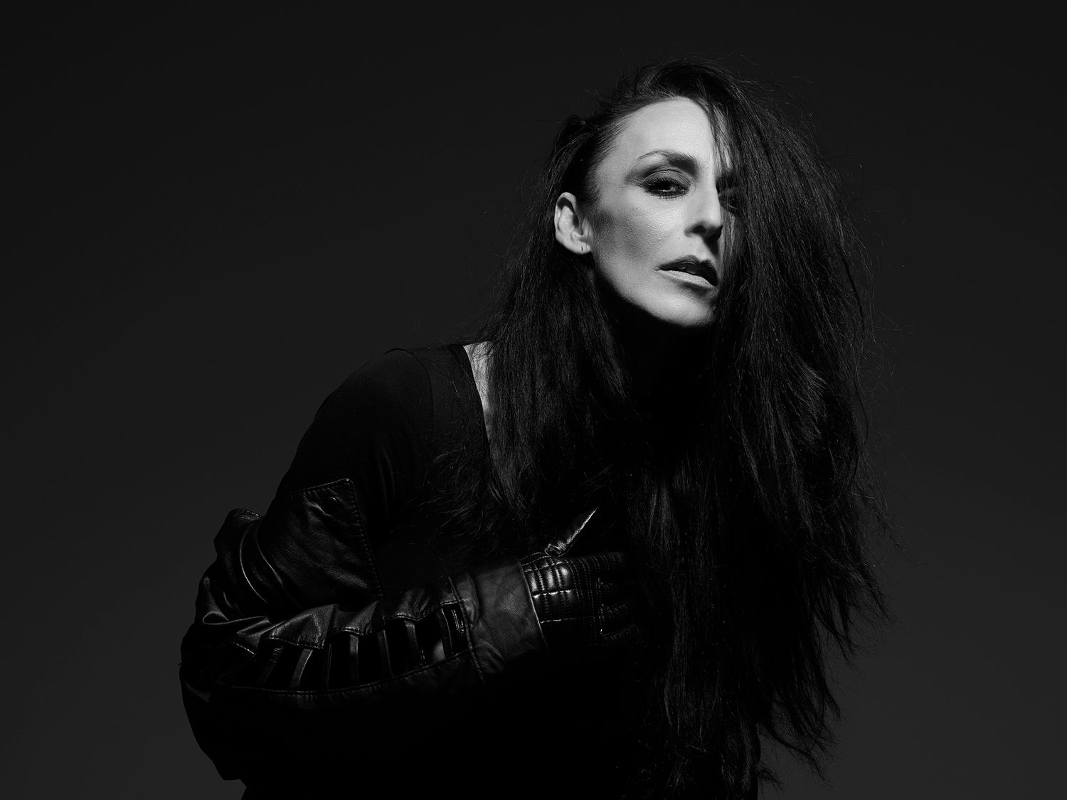 Black and white portrait of a woman with long dark hair in a leather jacket, posing dramatically against a dark background. Alba Hyseni