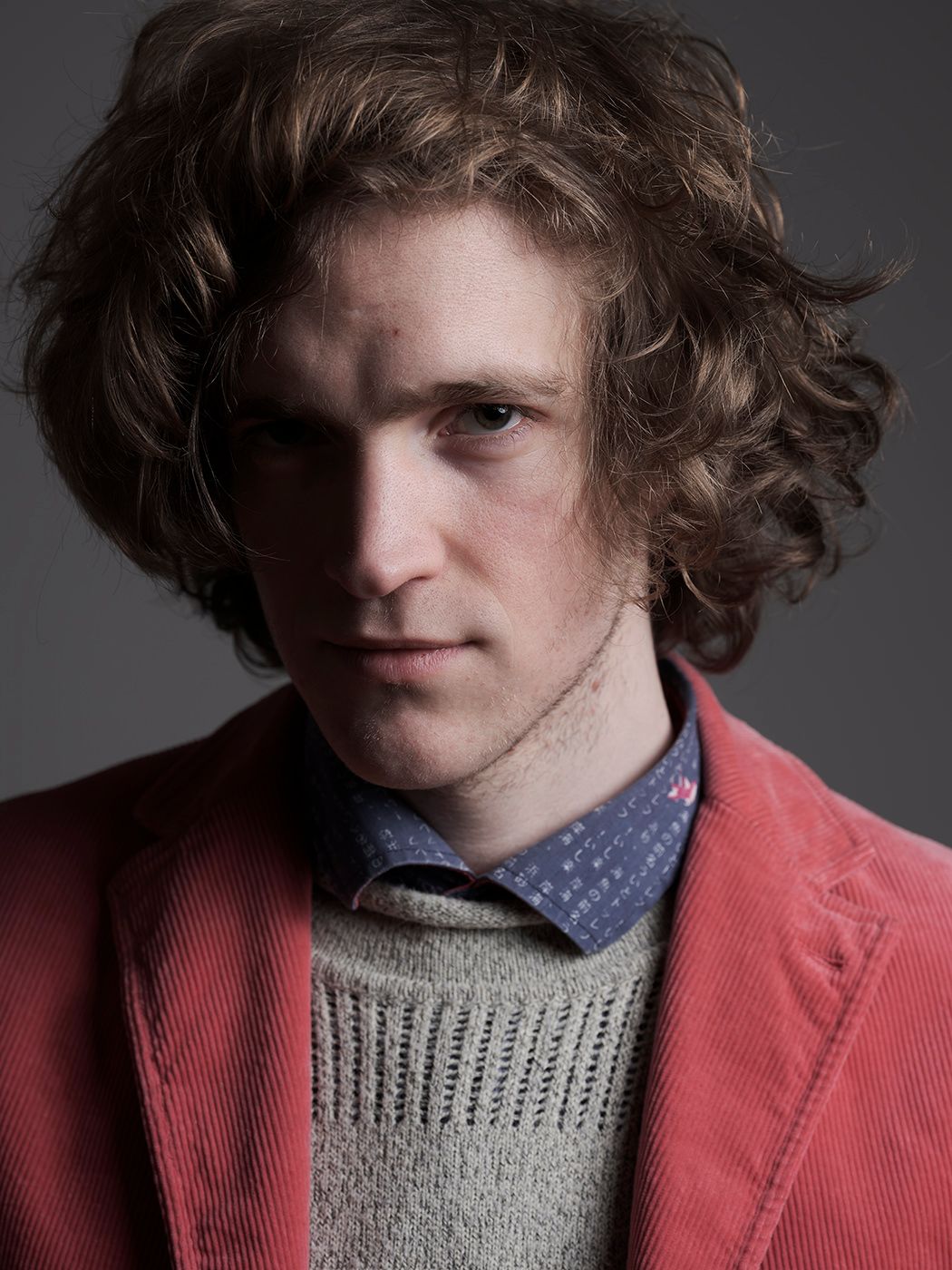 Young man with curly brown hair wearing a red blazer over a knit sweater and blue tie, serious expression. Gerlóczy Zsigmond