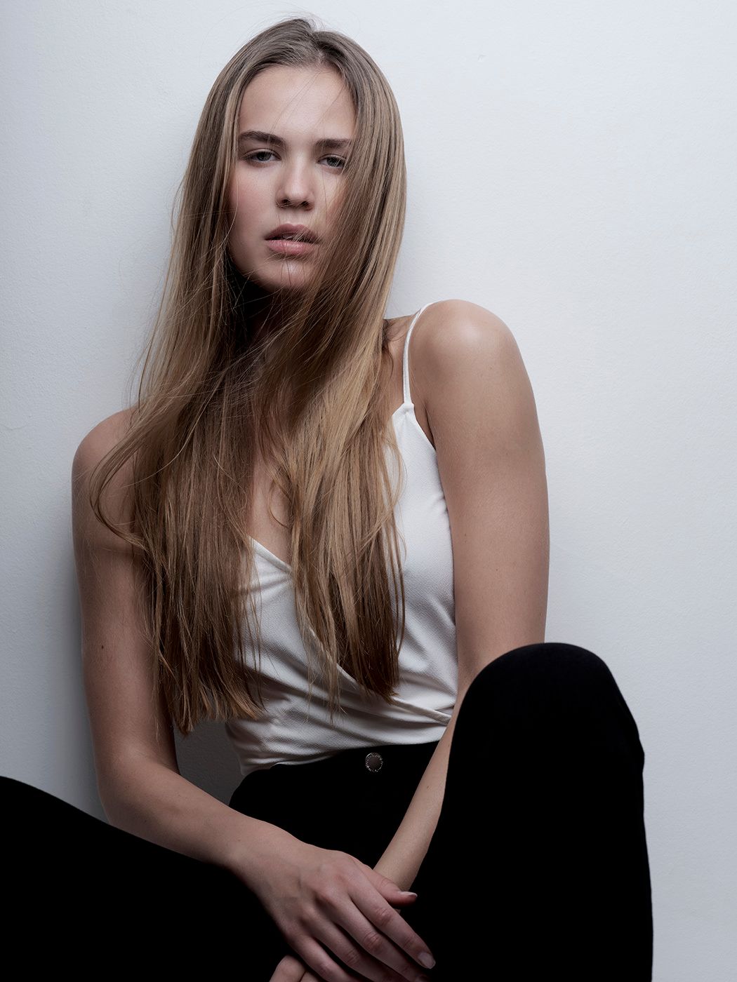 Young woman with long blonde hair in white tank top and black pants posing against a white wall.