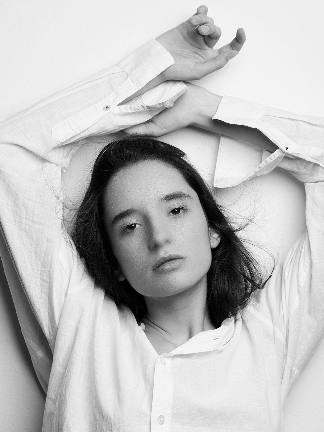 Black and white portrait of a young woman in a white shirt with arms raised above her head.