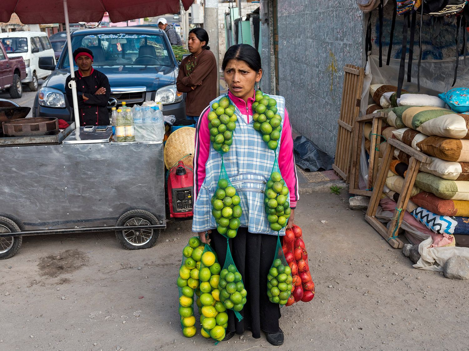 A woman vendor carrying large bunches of green limes in a busy outdoor market street.