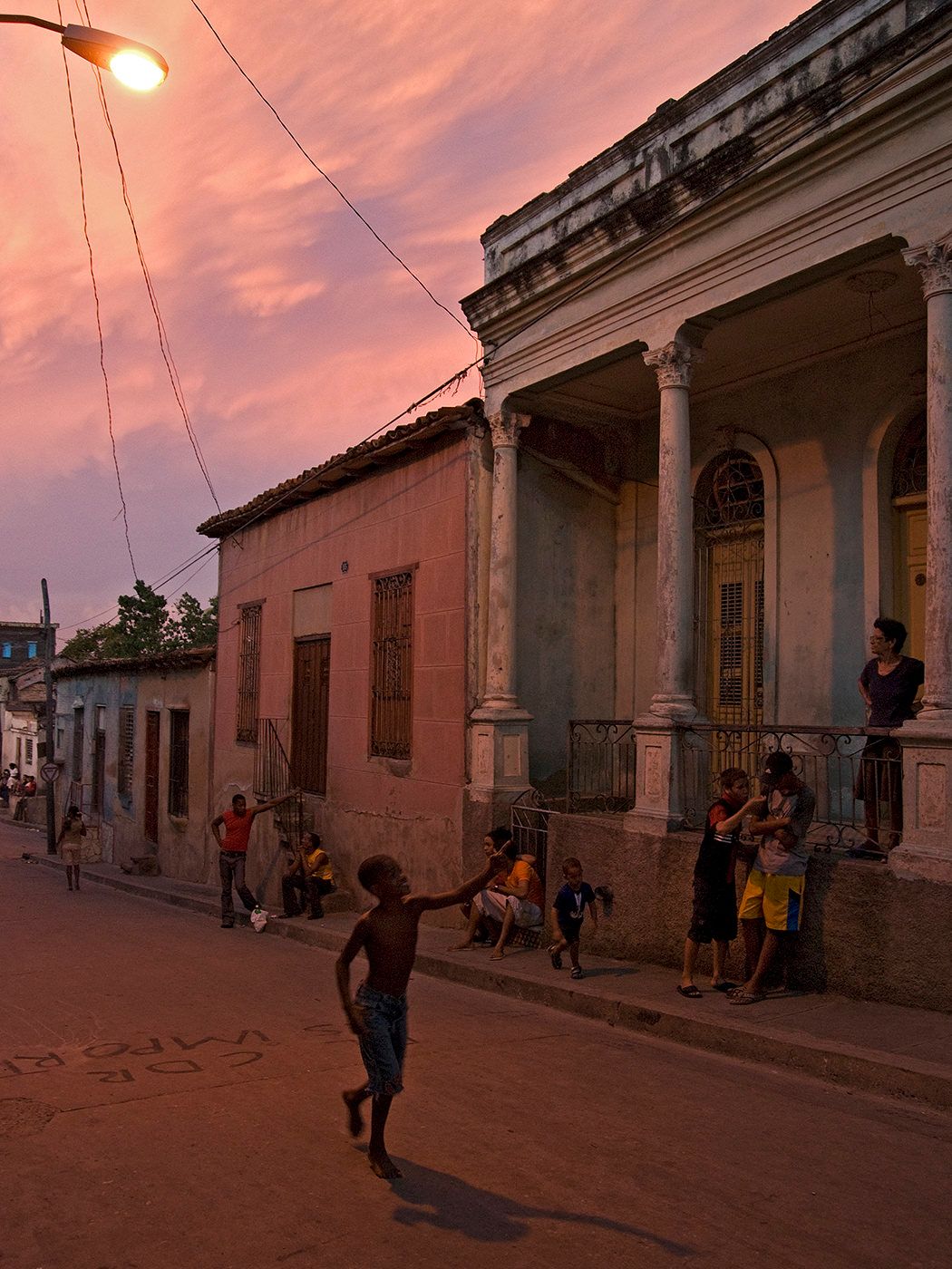 Children playing on a vibrant street at dusk in Cuba, with colorful colonial buildings and a dramatic pink sunset sky.