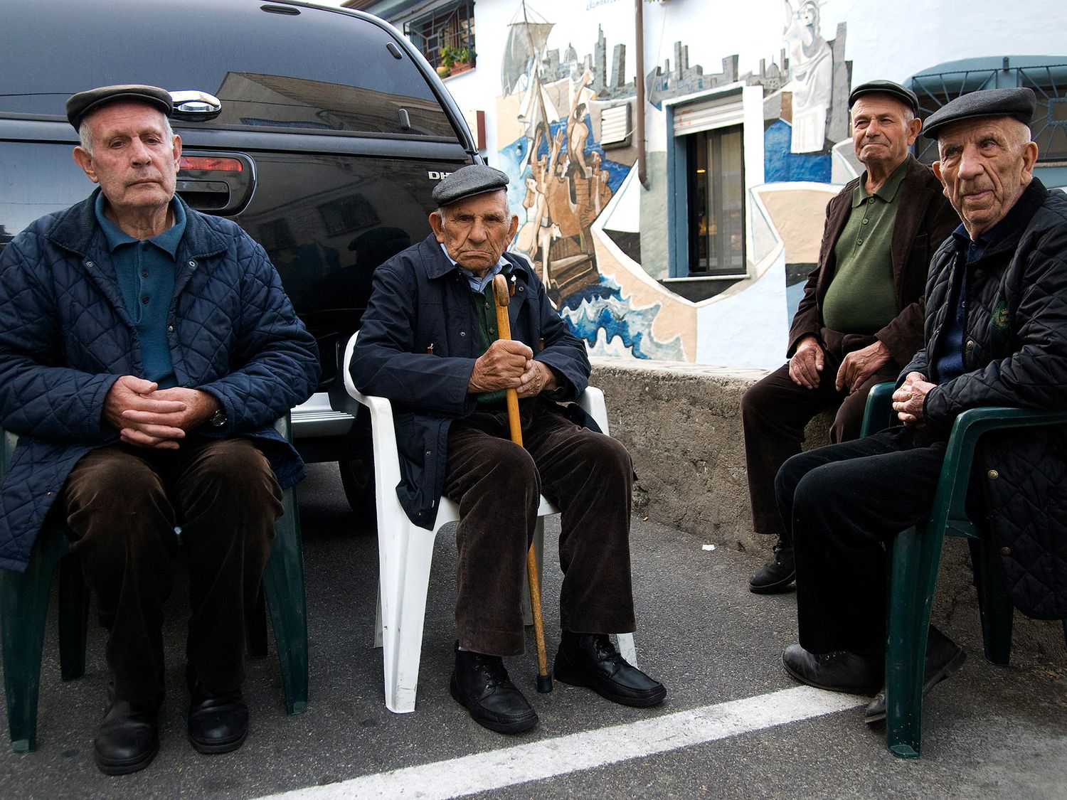Four elderly men sitting on chairs outdoors near a colorful mural, wearing caps and casual clothing.