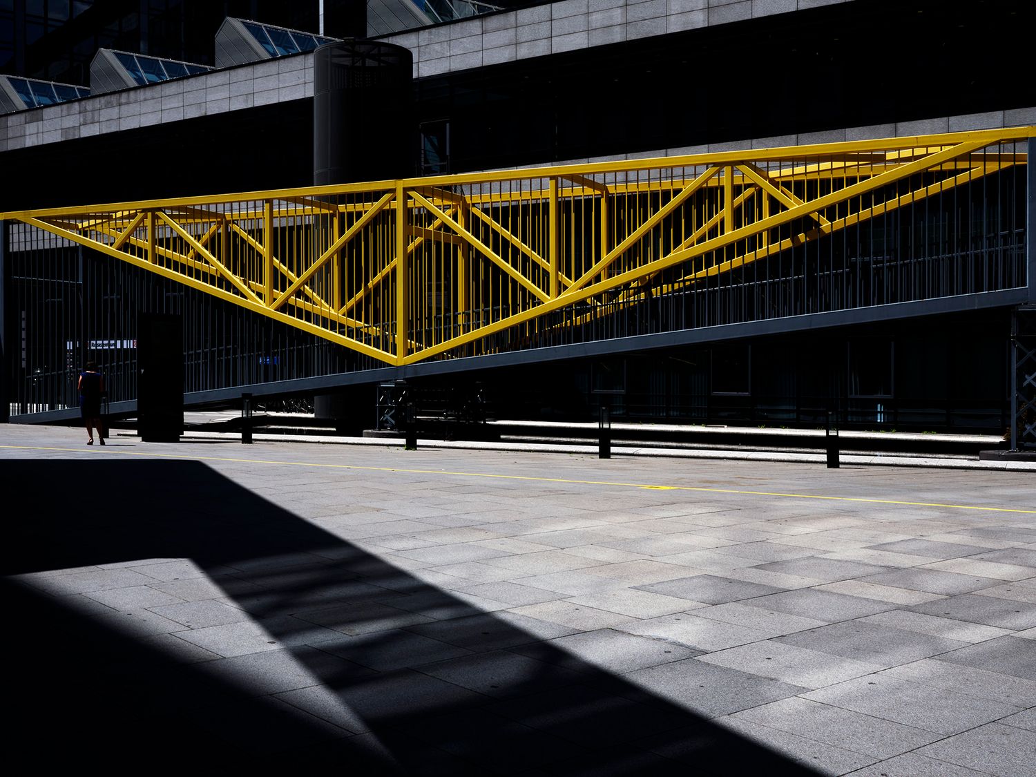 Yellow steel truss bridge structure in an urban plaza with dramatic shadows on grey pavement.