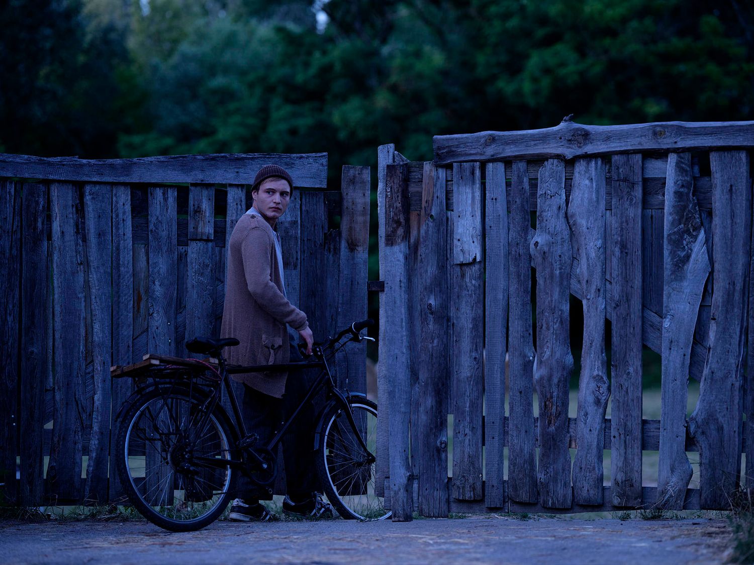 Young man with a bicycle standing by a weathered wooden fence in blue dusk light.
